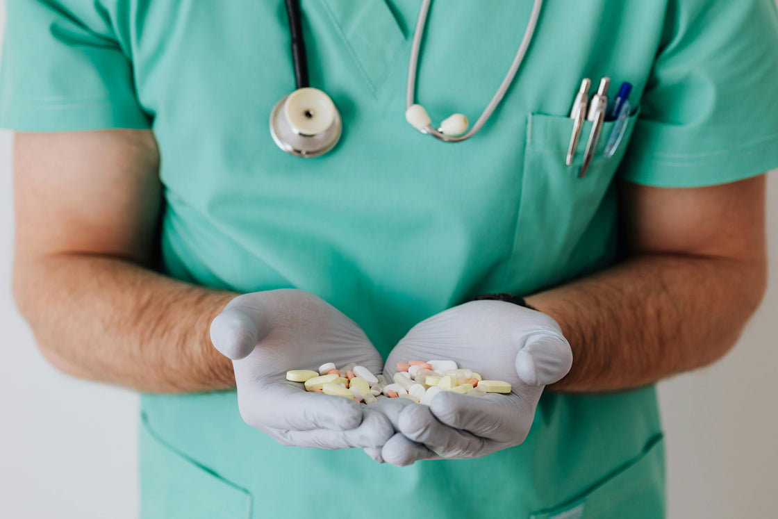 Person in green scrubs holding pills in gloved hands with a stethoscope around the neck.